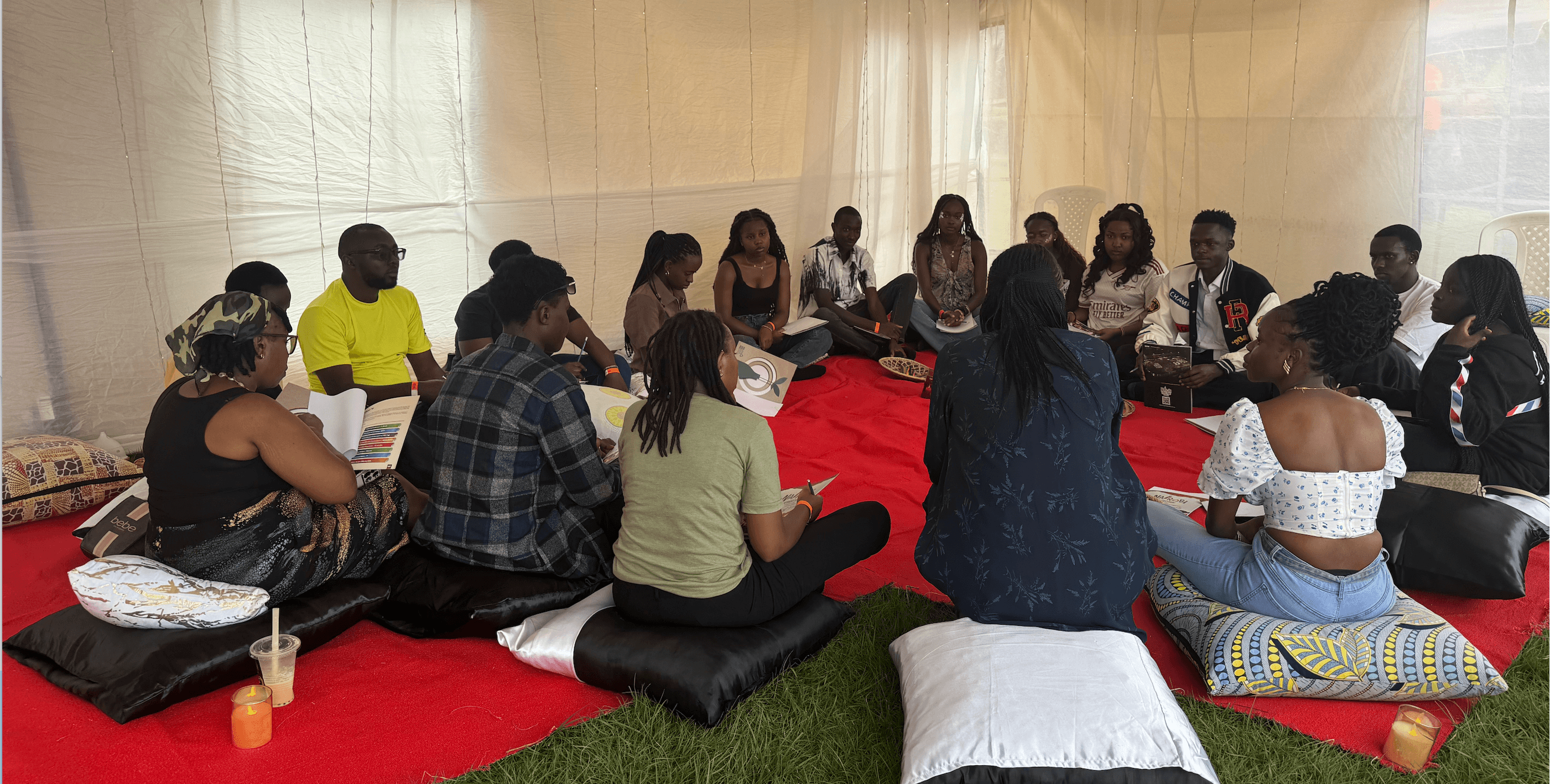 Participants seated in a circle on red mats in a tent, sharing a conversation